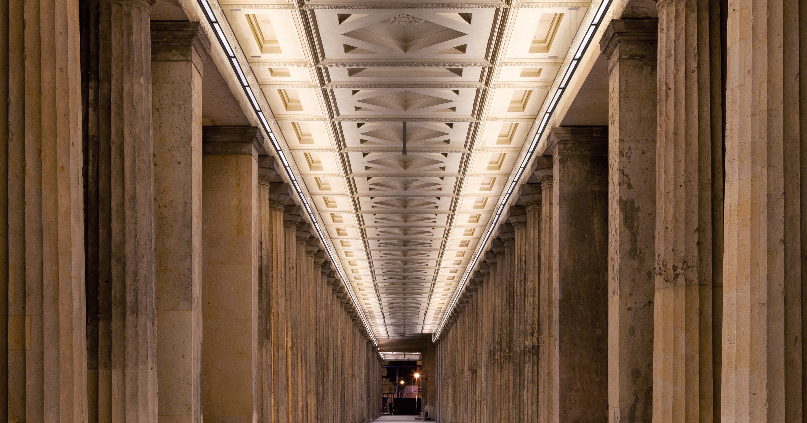 Une longue colonnade de pierre aux colonnes cannelées et au plafond à caissons éclairé, qui s’étire symétriquement vers une ouverture lointaine.